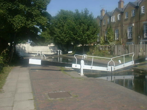 Paved area in foreground, lock, overhanging trees, brick building behind