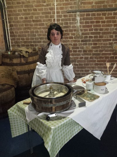 Lady in Edwardian costume stands behind a table on which sits a wooden churn and various utensils
