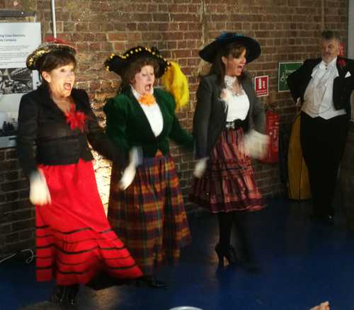 Four costumed performers sing, against brickwork background